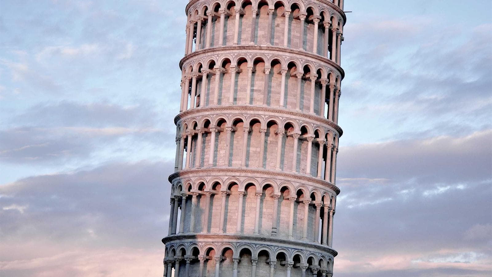 The Leaning Tower of Pisa is shown from an upward angle against a cloudy sky, highlighting its characteristic tilt and architectural details.